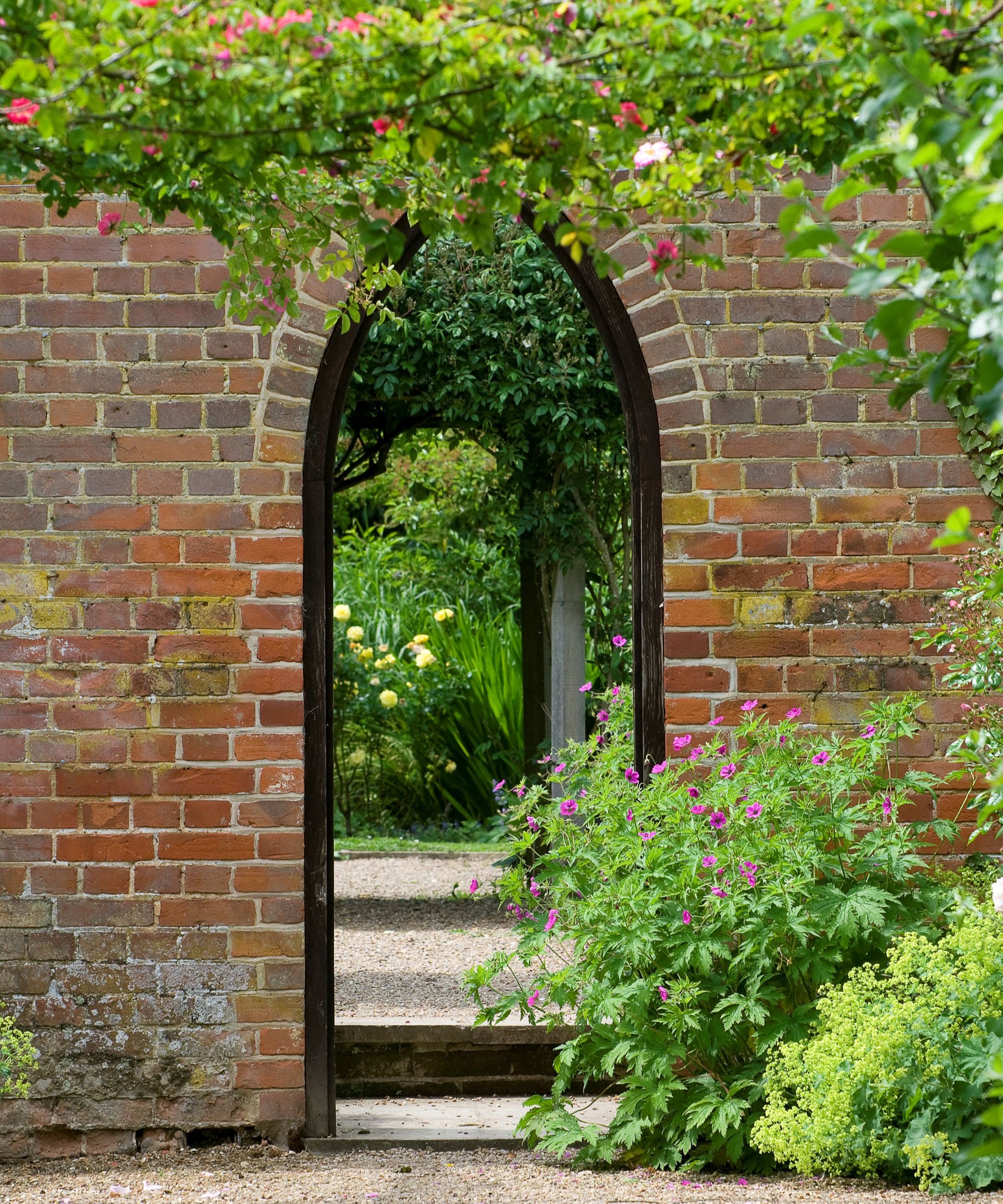An archway made from red brick with geranium plants in flower around it and gravel on the floor