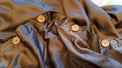 Four round, light-colored wooden buttons engraved with the words "PRETTY YOU HOME" are sewn onto the seam of a dark, silky draped fabric.