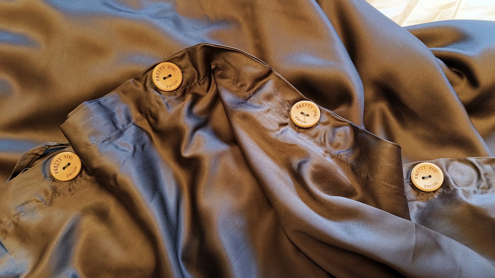 Four round, light-colored wooden buttons engraved with the words "PRETTY YOU HOME" are sewn onto the seam of a dark, silky draped fabric.
