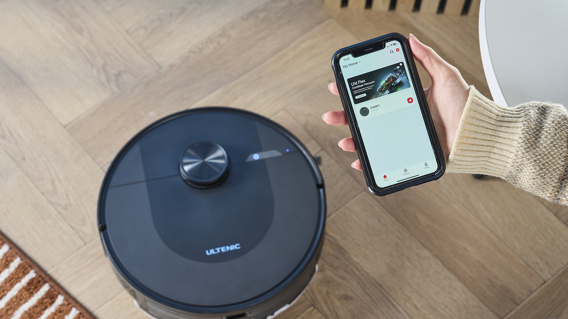 a rechargeable robot vacuum and mop in black by ultenic is photographed on a hardwood floor with a blue wall and a burnt orange terracotta and white striped rug