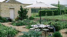 English country garden with flower beds, a brick outbuilding, a lawn looking out onto a field, and a round table with wrought iron bistro chairs and a parasol above 