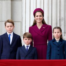 Prince William wears military uniform and Kate Middleton wears a dark red coat dress and matching hat as they stand with children George, Charlotte, and Louis on the Buckingham Palace balcony