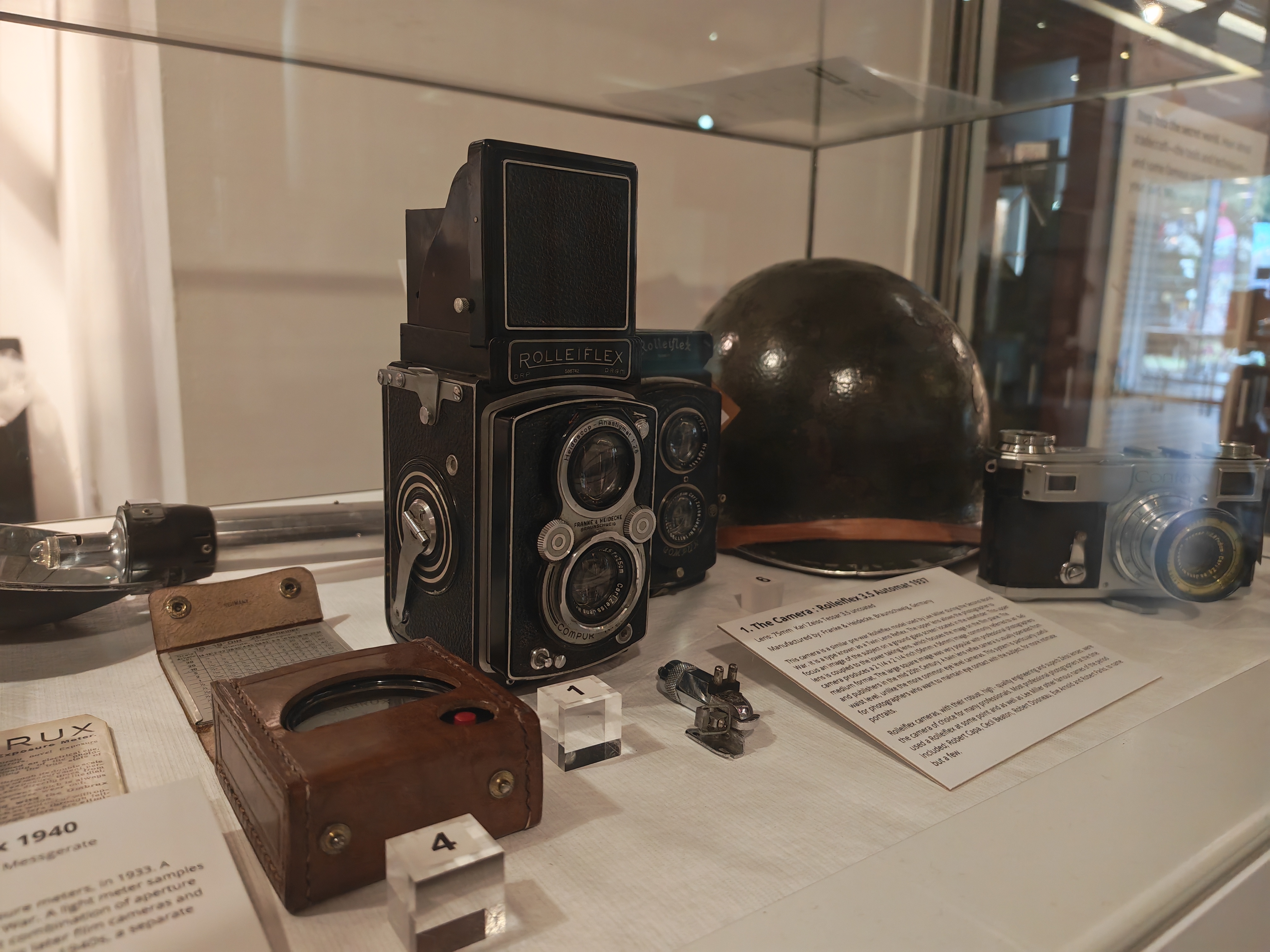 A museum display case containing Lee Miller's Rolleiflex camera, a US Army steel helmet, a compact camera and a light meter in a leather case, each labelled with exhibition notes.