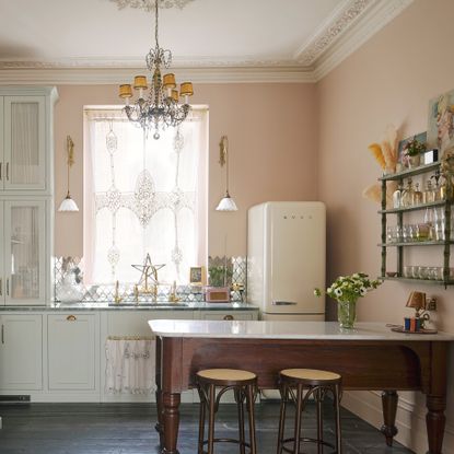 kitchen with pink walls and dark wood floors smeg fridge and lace covered window on the back wall with grey cabinetry below wooden farmhouse table with stools and open shelving unit