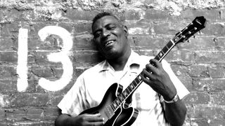 Howlin' Wolf standing against a brick wall holding a guitar