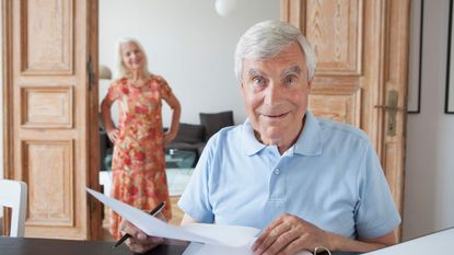 An older man looks up from papers with an enigmatic smile on his face as his wife stands in the background.