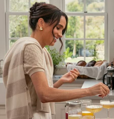 Meghan Markle wearing a cream sweater pouring fruit in a drink talking in a kitchen with Mindy Kaling