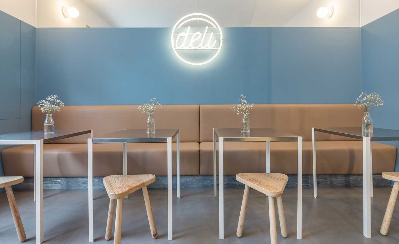 Interior view of the Deli restaurant. Painted in blue, with a neon &quot;Deli&quot; sign in the center, there is a sitting area in light brown that goes from wall to wall, in front of which are metal tables and short wooden stools.