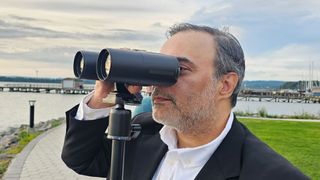 A man looking through the Unistellar Envision smart binoculars with a dock in the background.