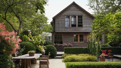 A lush, tiered backyard garden with manicured hedges, stone pathways, and a large outdoor wooden dining table. In the background stands a weathered two-story grey shingle house surrounded by mature trees.