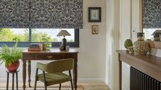 A small wooden home office desk with a green dining-style arm chair pulled up to it. A large blue and white floral roman blind on the window above. 