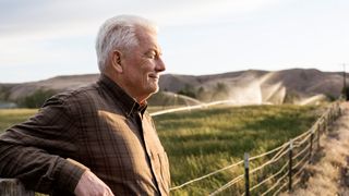 Senior man leans back on wooden gate against country landscape, aging in place successfully