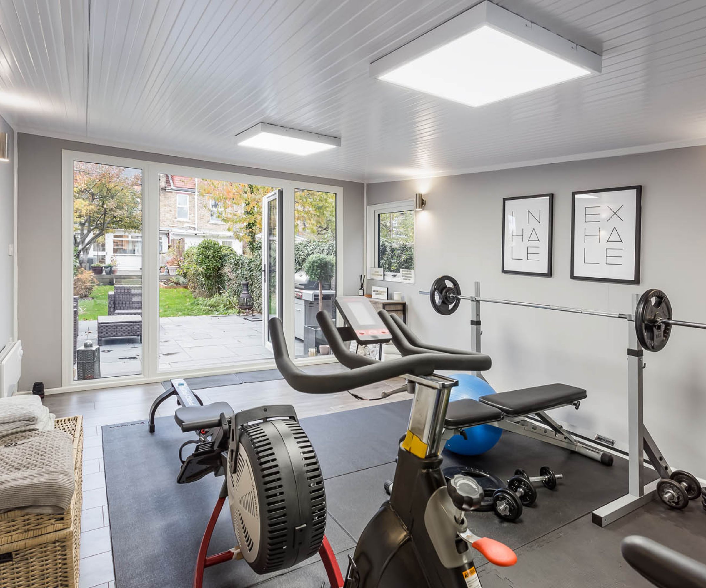 Grey home gym with spin bike, rower, weight bench and barbell rack, glass doors open to garden.