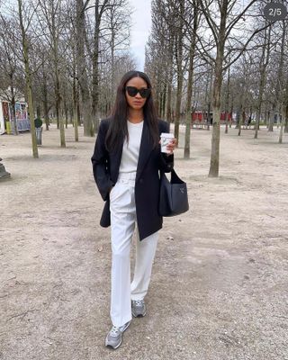 British influencer Lorna Humphrey of Symphony of Silk poses in a park with trees wearing black oversize sunglasses, black blazer, white t-shirt, white trousers, and gray New Balance sneakers