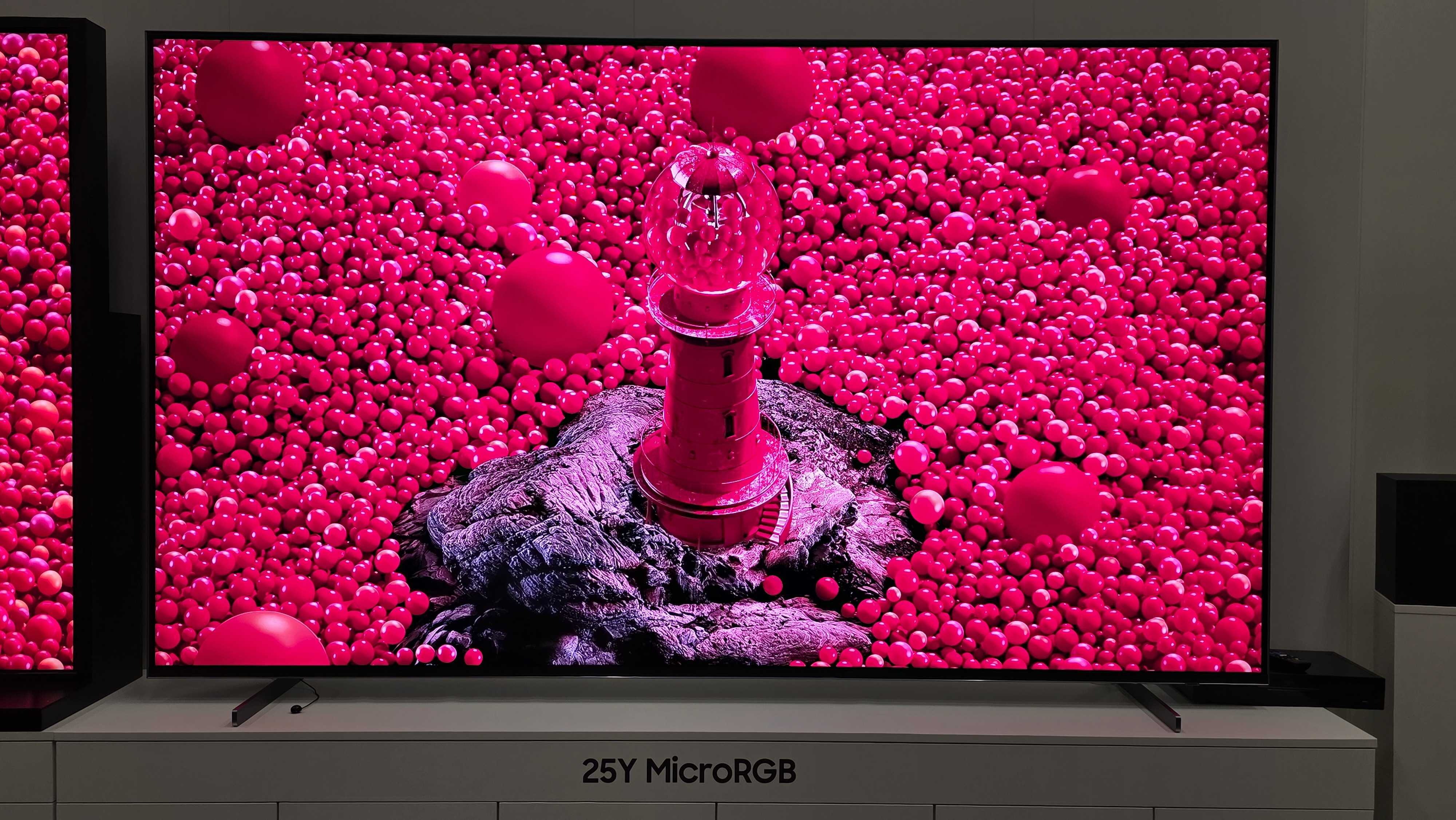 A 115-inch Samsung RGB LED TV on a white stand with colourful demo content on screen featuring the Statue of Liberty and a hot air balloon made of roses.