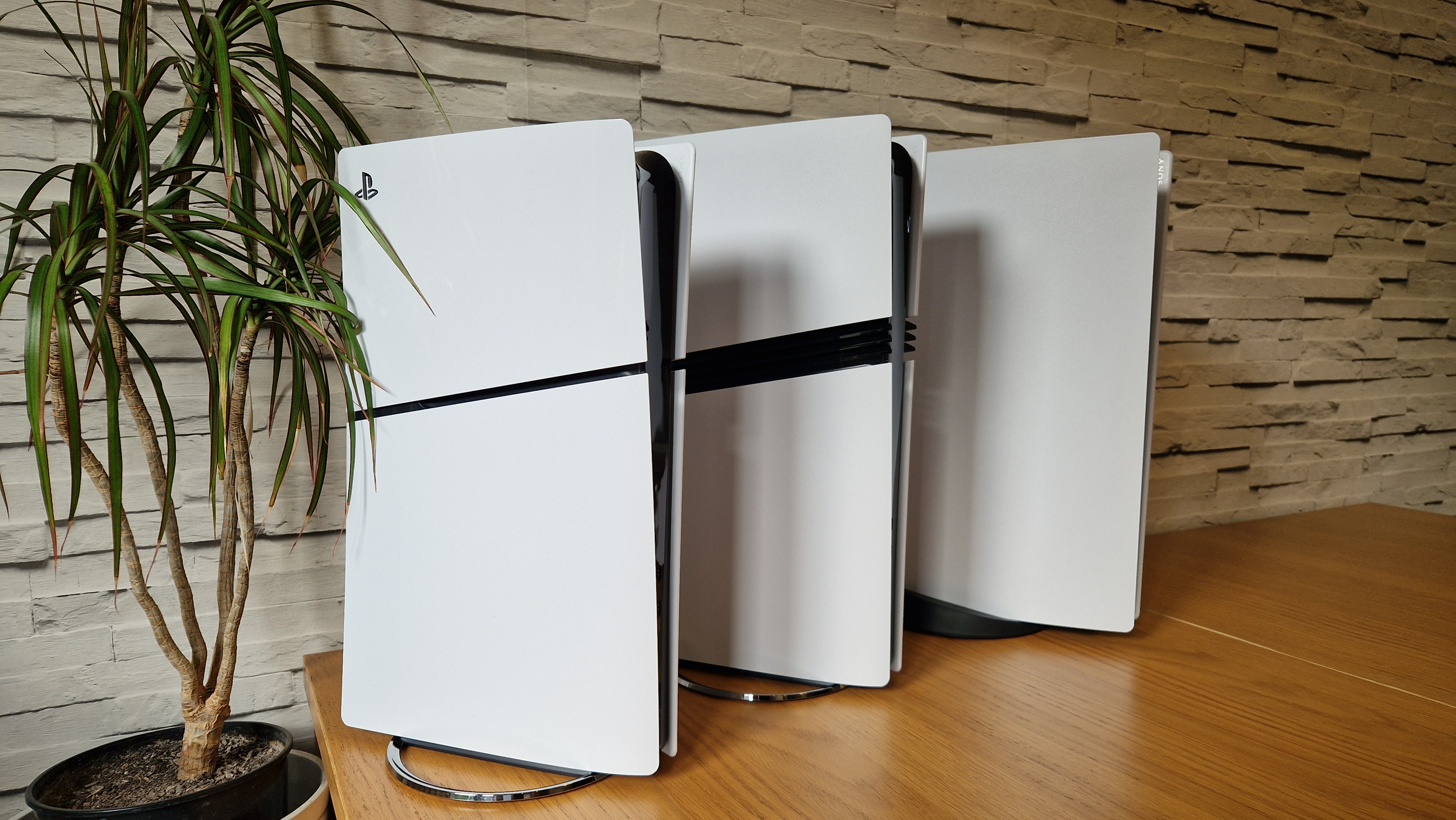 The PS5 Slim next to a PS5 Pro and launch model PS5 on a wooden table with a white brick background and next to a vase of flowers