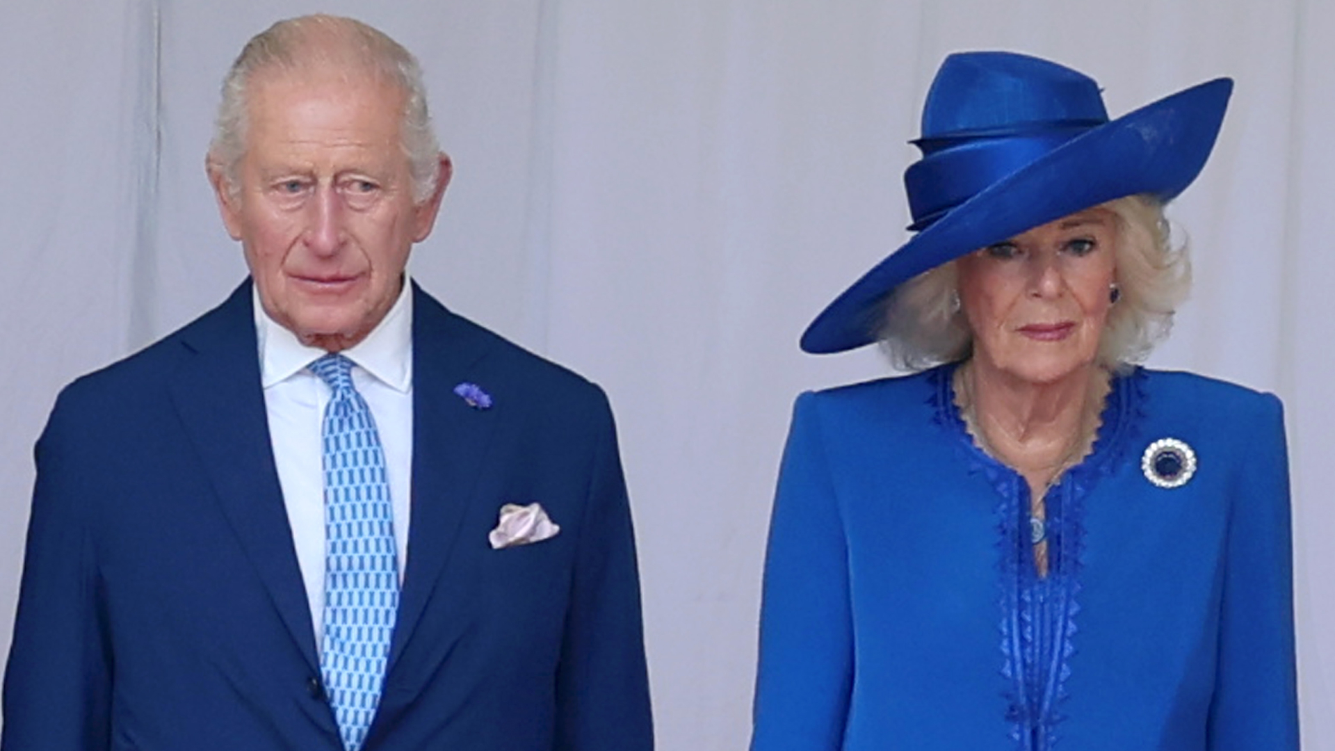 King Charles wears a navy suit with a light blue tie while Queen Camilla wears a royal blue dress with a matching hat