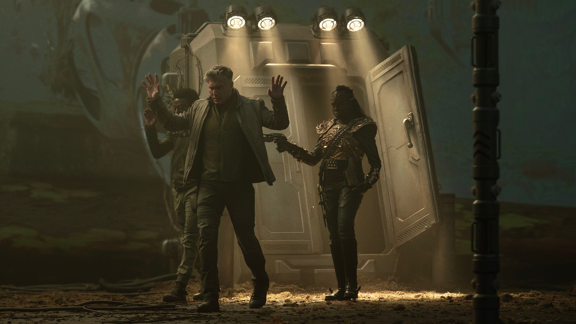 An angry female Klingon is pointing a gun at two man. Both men are holding their hands up in surrender and walking where they are directed. L to R Babs Olusanmokun as Dr. MíBenga, Anson Mount as Capt. Pike, and Christine Horn as Bytha in season 3, Episode 3 of Strange New Worlds streaming on Paramount+.