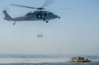 a helicopter in mid-air with a basket underneath. at right is a raft with several sailors on board, floating in the ocean