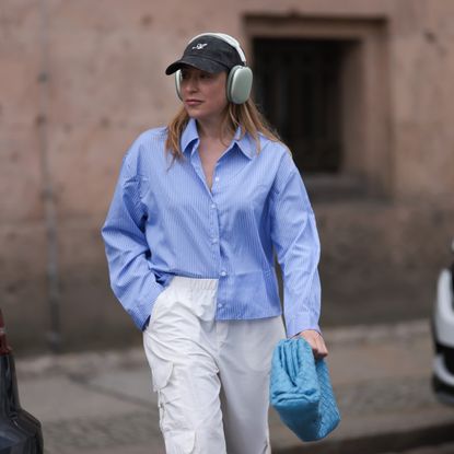 Sonia Lyson wearing Zara hose, Storets blue and white striped long sleeve buttoned cropped flannel, Bottega Veneta blue leather pouch bag, Axel Arigato dark grey logo cap, Apple AirPods Max olive color headphones on June 28, 2023 in Berlin, Germany.