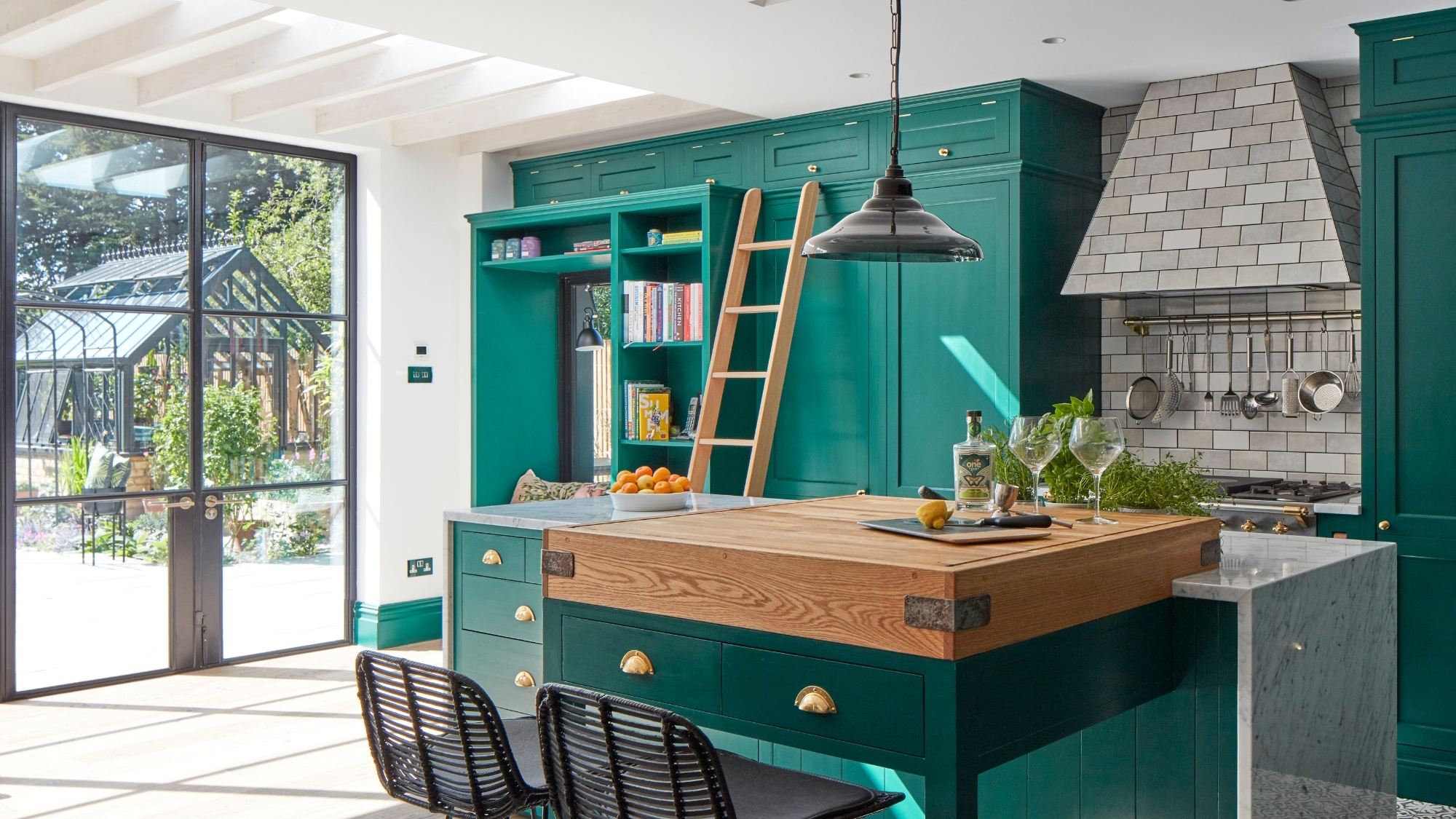 Green kitchen island with oak chopping-block top, ladder, pendant light, glazed doors to garden beyond.