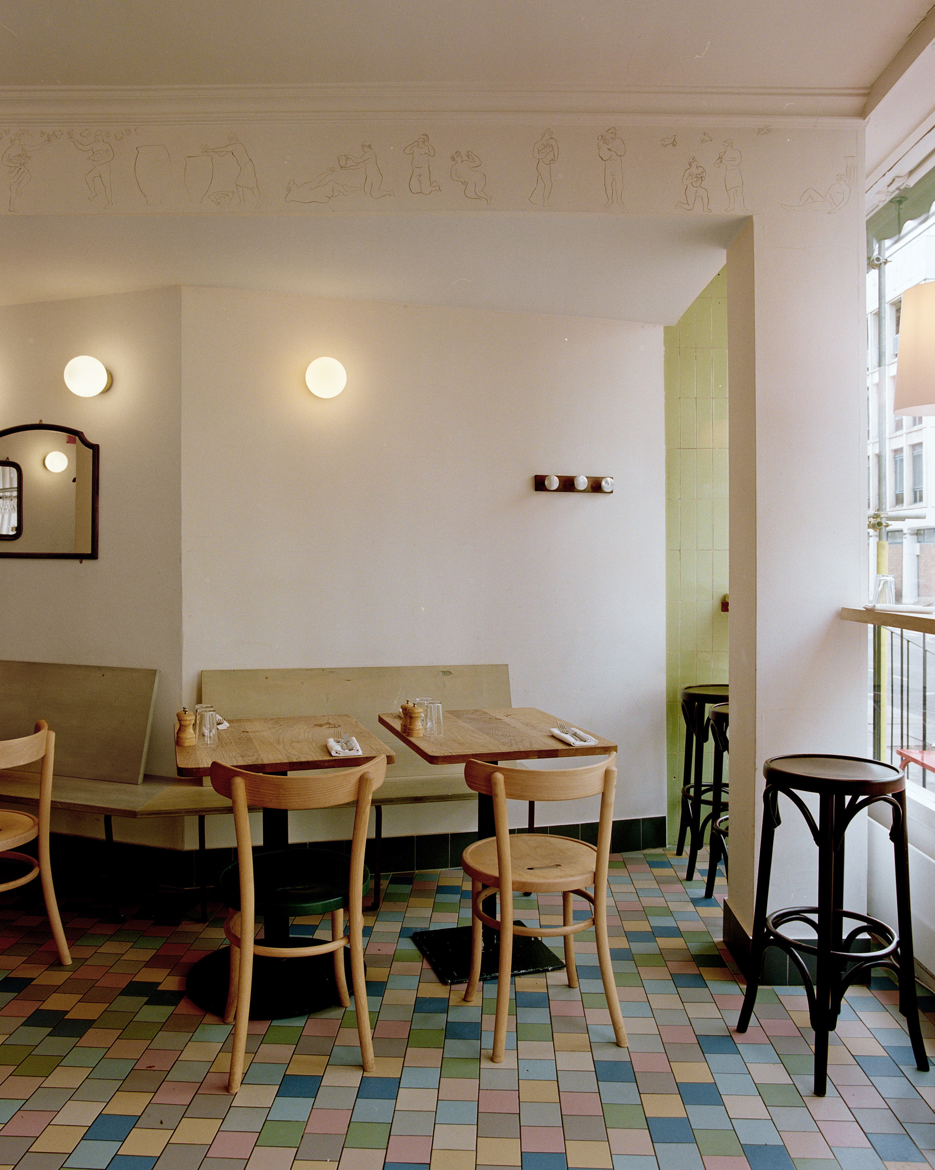 An Art Deco-style cafe and restaurant with pastel checkered floors in green, pink, blue, grey, yellow, and terracotta tones, celery green walls, round lighting, and hand-drawn frieze decor.