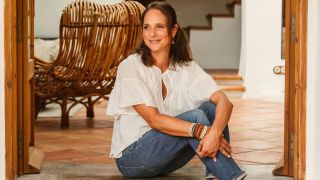 A woman with brunette hair, a white loose blouse, and blue denim, with leather bracelets in brown and blue and cream tones, sits on the cement stairs of her outdoor patio with terracotta flooring.