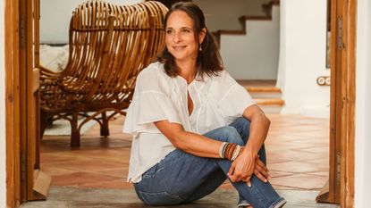 A woman with brunette hair, a white loose blouse, and blue denim, with leather bracelets in brown and blue and cream tones, sits on the cement stairs of her outdoor patio with terracotta flooring.