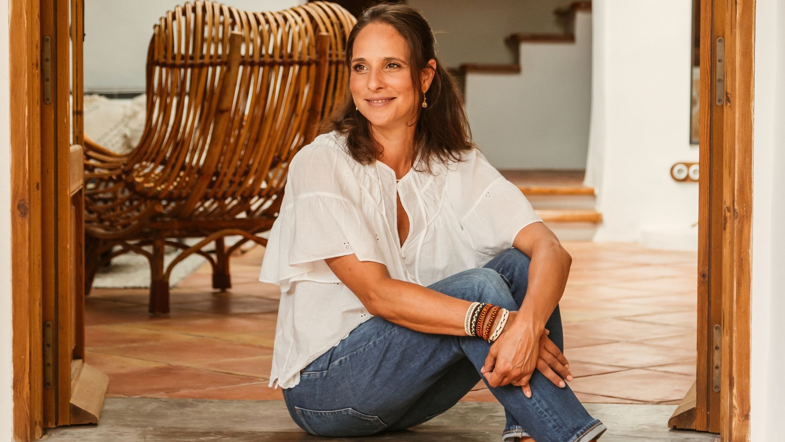 A woman with brunette hair, a white loose blouse, and blue denim, with leather bracelets in brown and blue and cream tones, sits on the cement stairs of her outdoor patio with terracotta flooring.