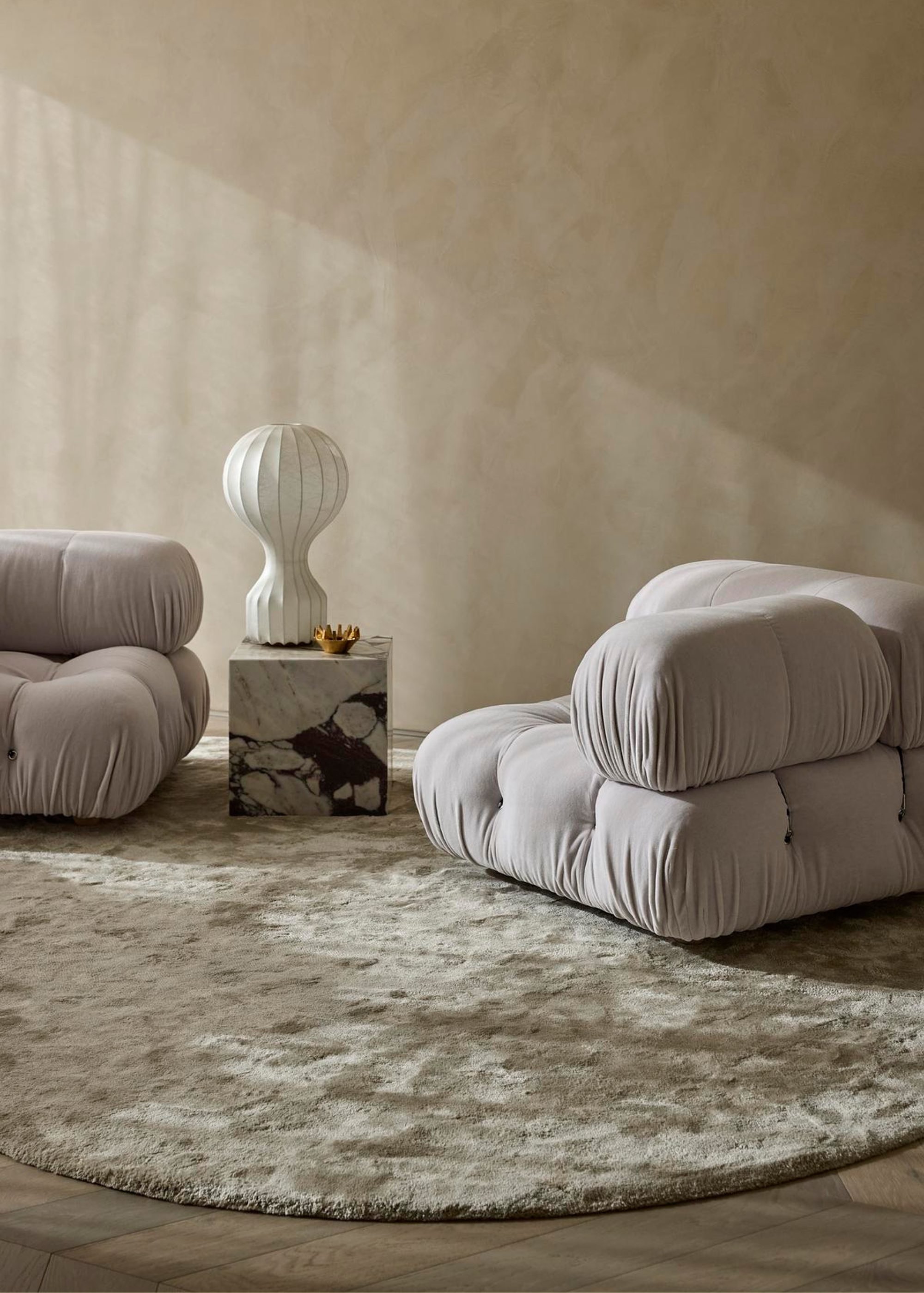A beige room with sofa chairs, a stone side table, a paper lamp, and a round fuzzy rug