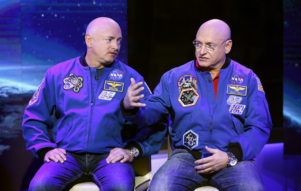 Scott and Mark Kelly sit side by side wearing a blue NASA jacket and jeans