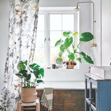 White bathroom with a bath with a shower over it, and a floral shower curtain surrounded by plants