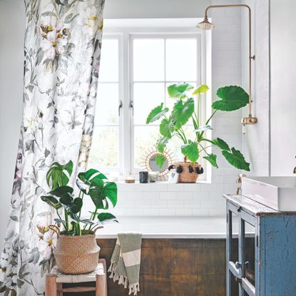 White bathroom with a bath with a shower over it, and a floral shower curtain surrounded by plants