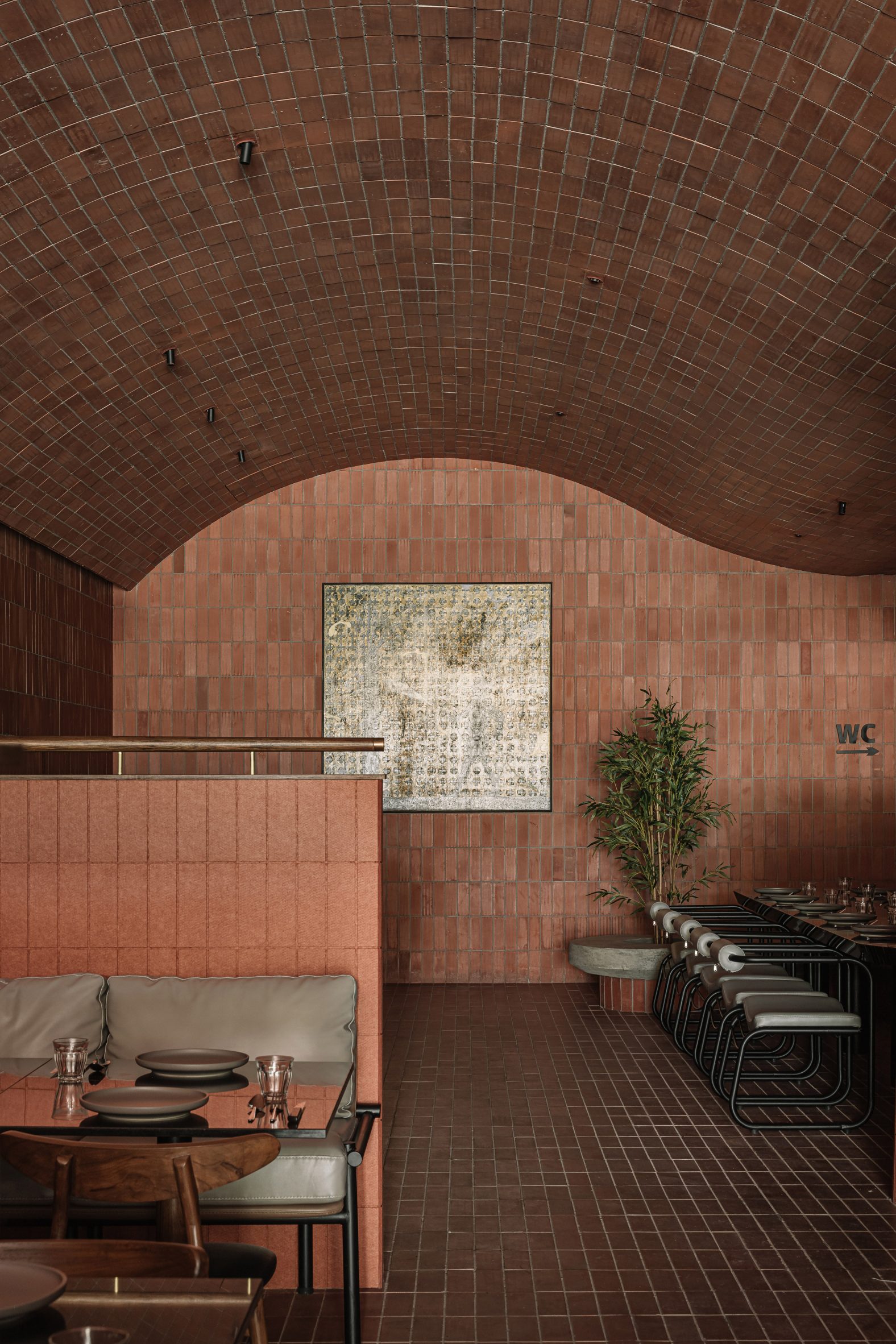 A brick-clad restaurant with terracotta walls, floors, and room dividers, neutral wrought iron and pale beige leather furniture, wooden tables, plants, and minimal decor boasts a wave-shaped ceiling.