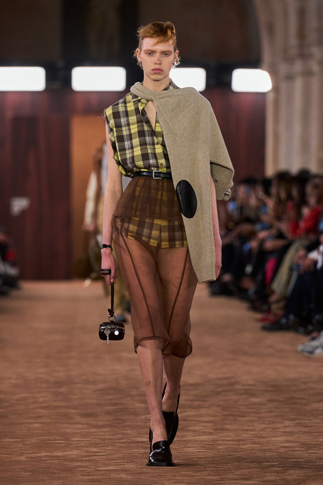 Layering staples for spring 2026 are shown in a photo from Acne's runway show, with a model walking while wearing a tan sweater tied around the neck styled with a bright yellow flannel shirt thats tucked into a brown sheer skirt worn with black loaders, a black belt, and a brown mini bag
