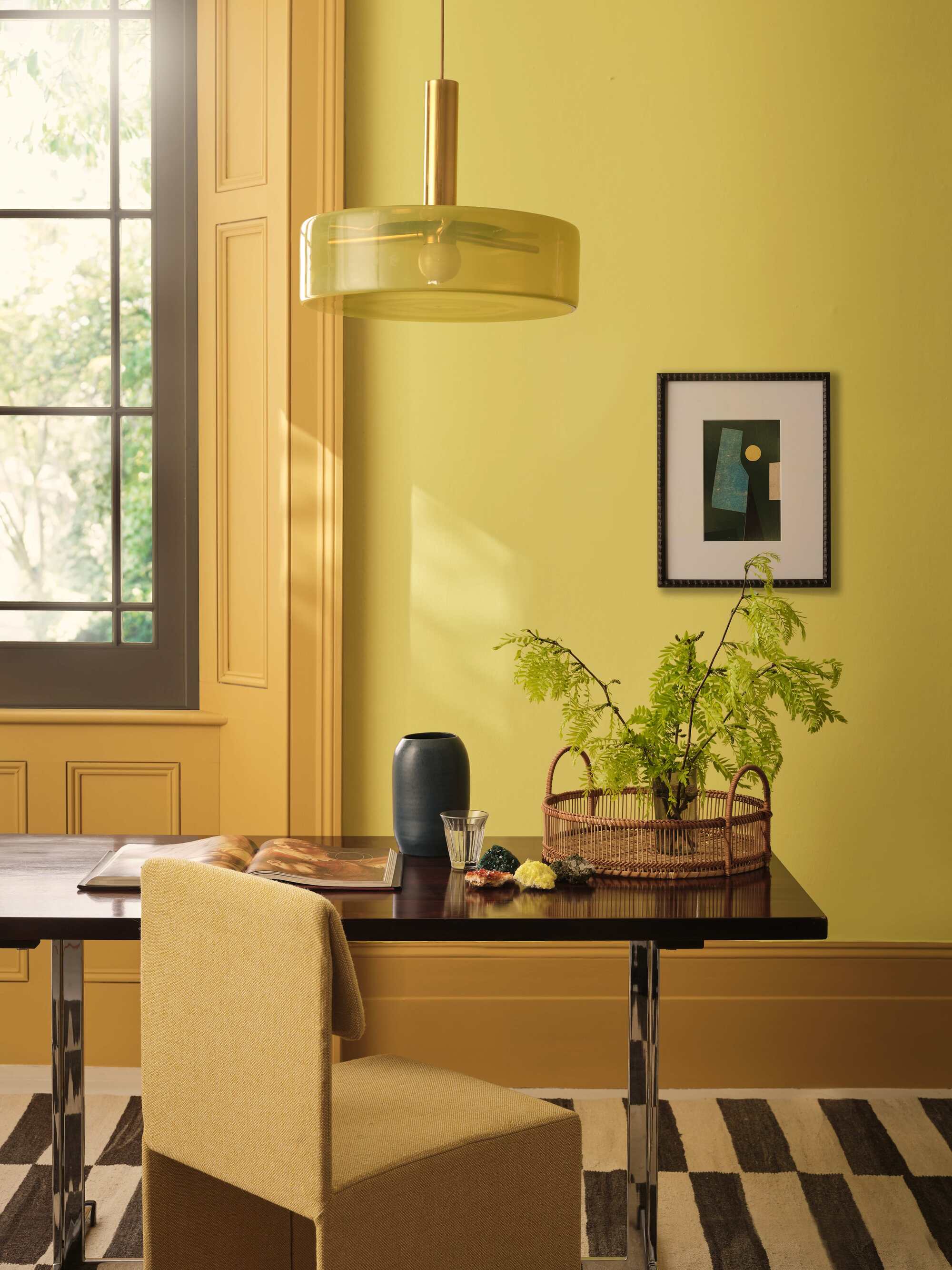 yellow home office with wall molding around windows, artwork on wall, striped black and white rug with desk styled with magazine, vase, and glass with foliage, and yellow upholstered chair