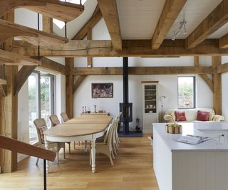 A compact oak frame home with lots of exposed beams and a spiral oak staircase to the left of the shot. The main room view is an open plan living room, dining room and kitchen. The interiors are neutral but classic and there is a log burning stove on the far wall.