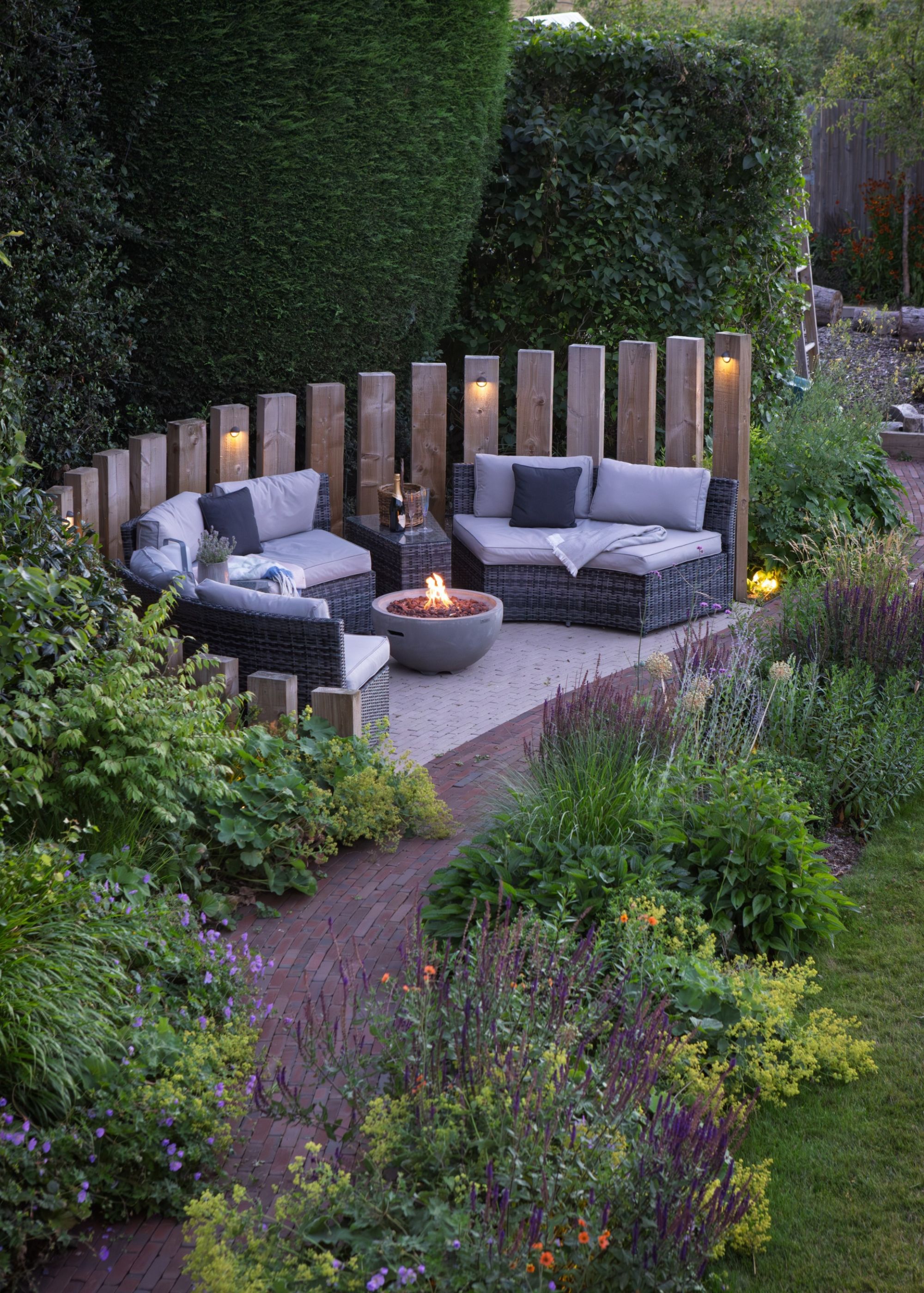 A zoned outdoor seating area with a fire pit in a calming garden surrounded by flower borders planting and large shaped trees