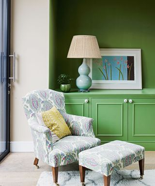 Corner of living room by window, green painted wall and cabinetry, upholstered lounge chair with patterned fabric, dark wooden legs, matching footstool