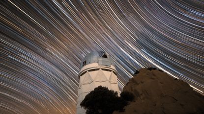 A long exposure photo shows the series of white star streaks across the night sky above a circular observatory.