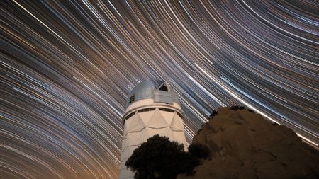A long exposure photo shows the series of white star streaks across the night sky above a circular observatory.