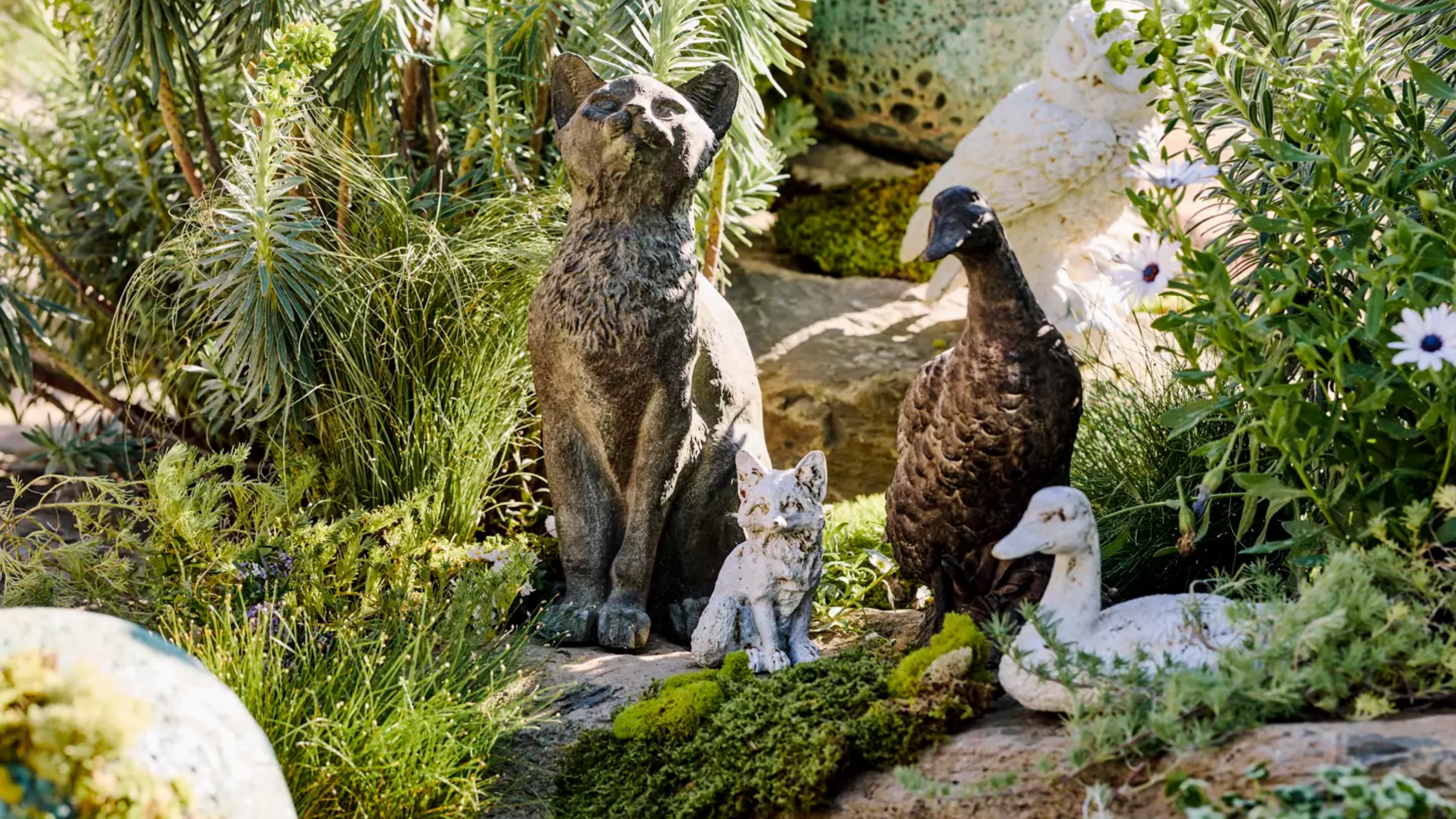 a collection of garden statues including cats and ducks surrounded by plant life