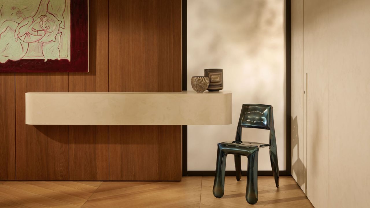 Image of a minimalist entryway that has beige walls, wood floors, and the back wall is wood paneled. There is a large white marble shelf attached to the wall and a metal chair to the right of the shelf.