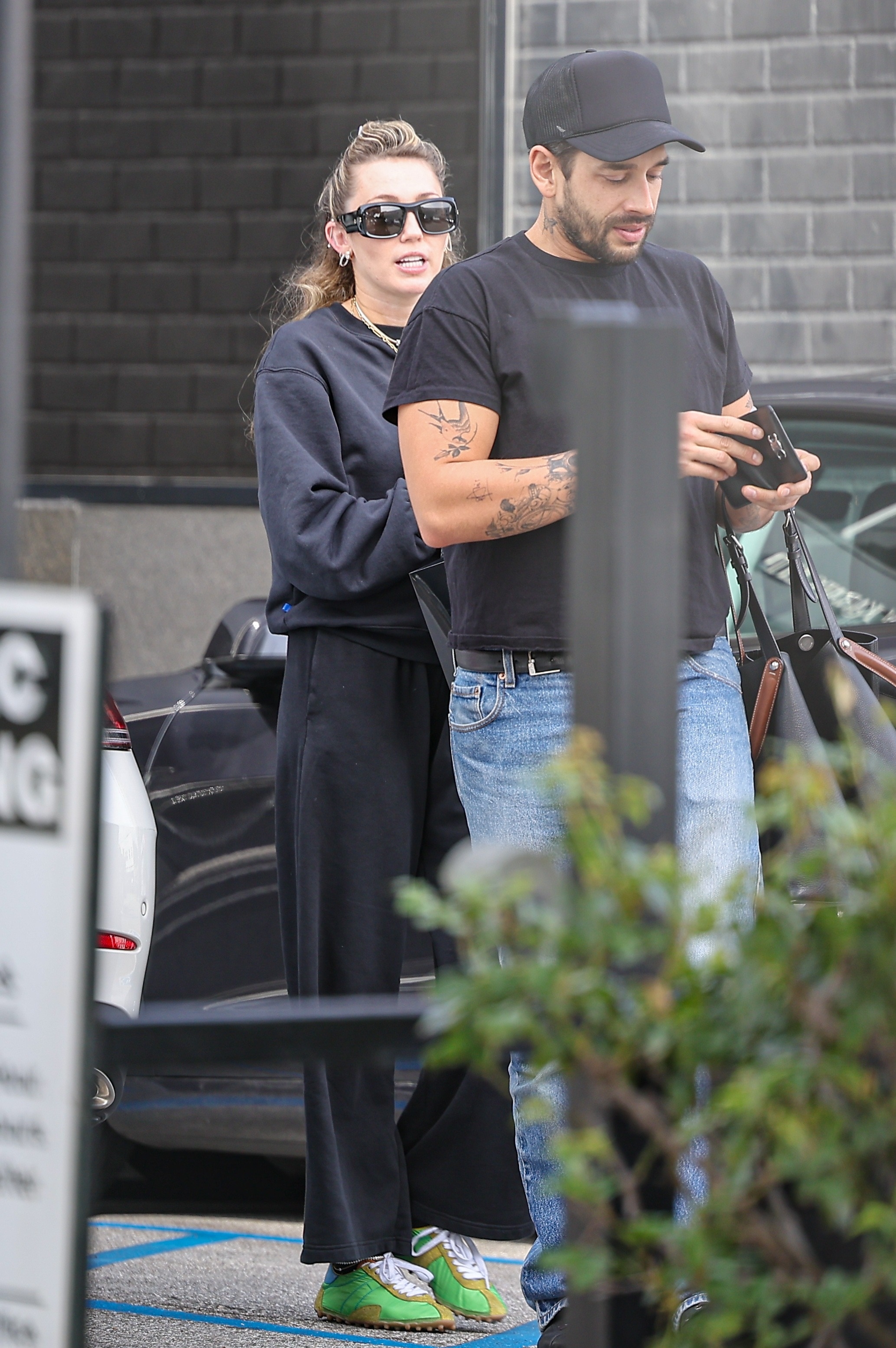 Miley Cyrus wears a black top and black track pants with green trainers and black sunglasses.