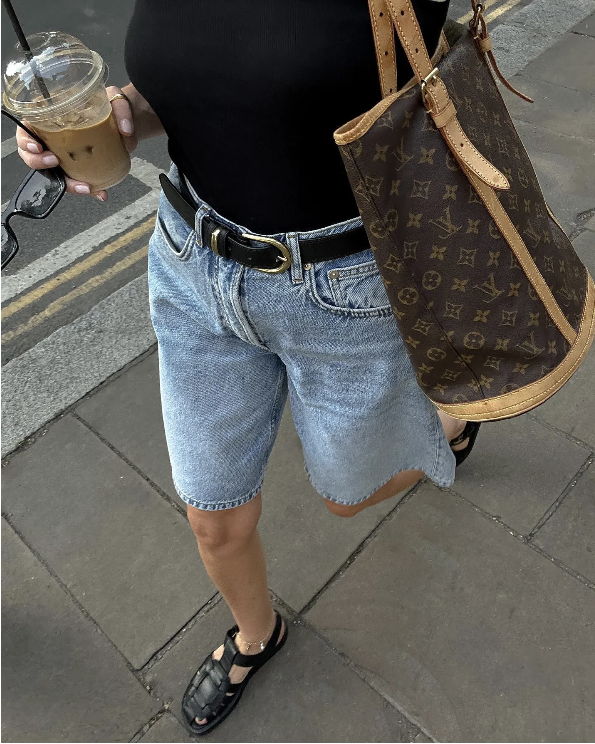 Stylist Francesca Saffari wears blue denim jorts with black fisherman sandals. She pairs the outfit with a black belt, a matching black tee and a Louis Vuitton shoulder bag. In the recent image taken from her Instagram, you can't see her face, just her iced coffee and the pavement that she's walking on