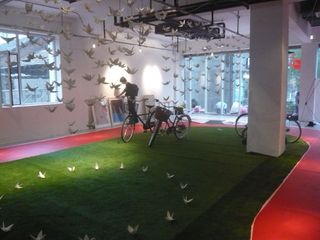 Artificial grass has been laid on the floor of an old computer factory. Origami cranes sit in a circle on the grass, and many more are suspended from the ceiling.