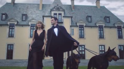 Taylor Swift - Blank Space video still, wearing black dress, walking with man in tux, holding a Doberman on a leash