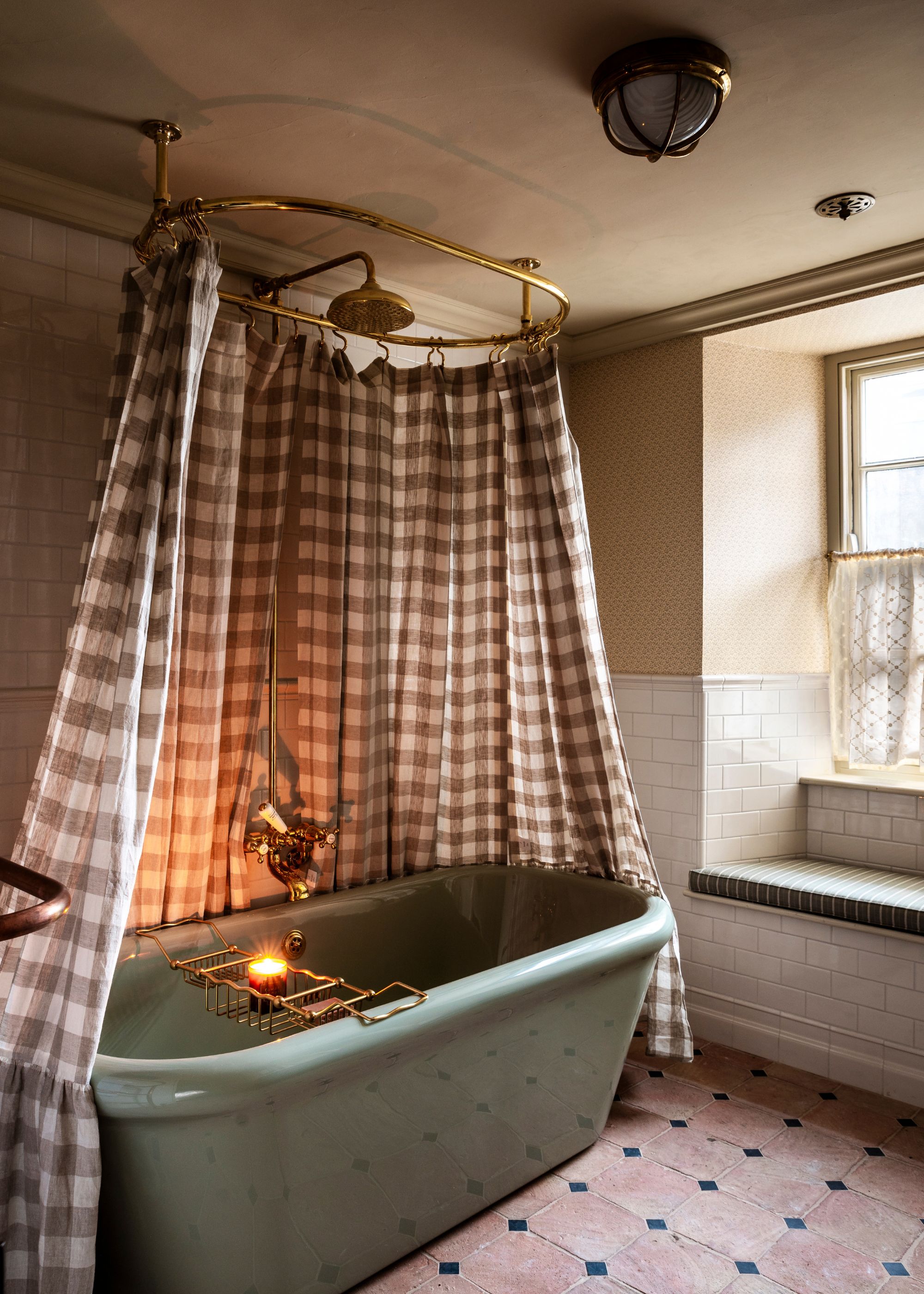 A bathroom with neutral floral wallpaper on the upper walls. white tiles on the lower walls, terracotta floor tiles, and a bath in the middle of the room with a gingham shower curtain.