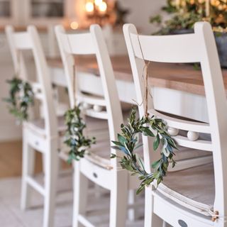 a close up of some white dining chairs with decorative festive wreaths hung from each