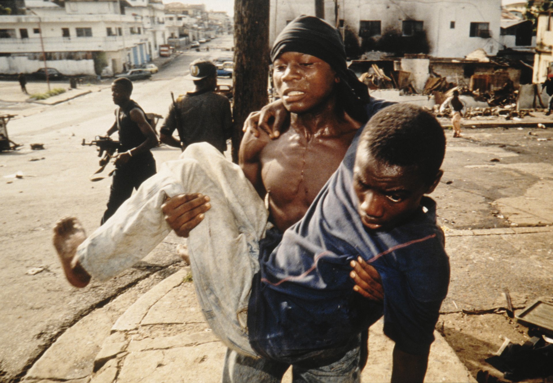 01 January, 1996 An NPFL Patriotic Front fighter carries a wounded comrade to safety. In April and May excessive violence flares up. The fiercest fighting in the country's seven-year civil strife claimed some 1,500 lives, forcing most aid organizations to evacuate their workers and almost half the population to flee their homes. The situation was sparked off by the refusal to surrender of a dismissed minister and warlord supported by the Krahn ethnic group.